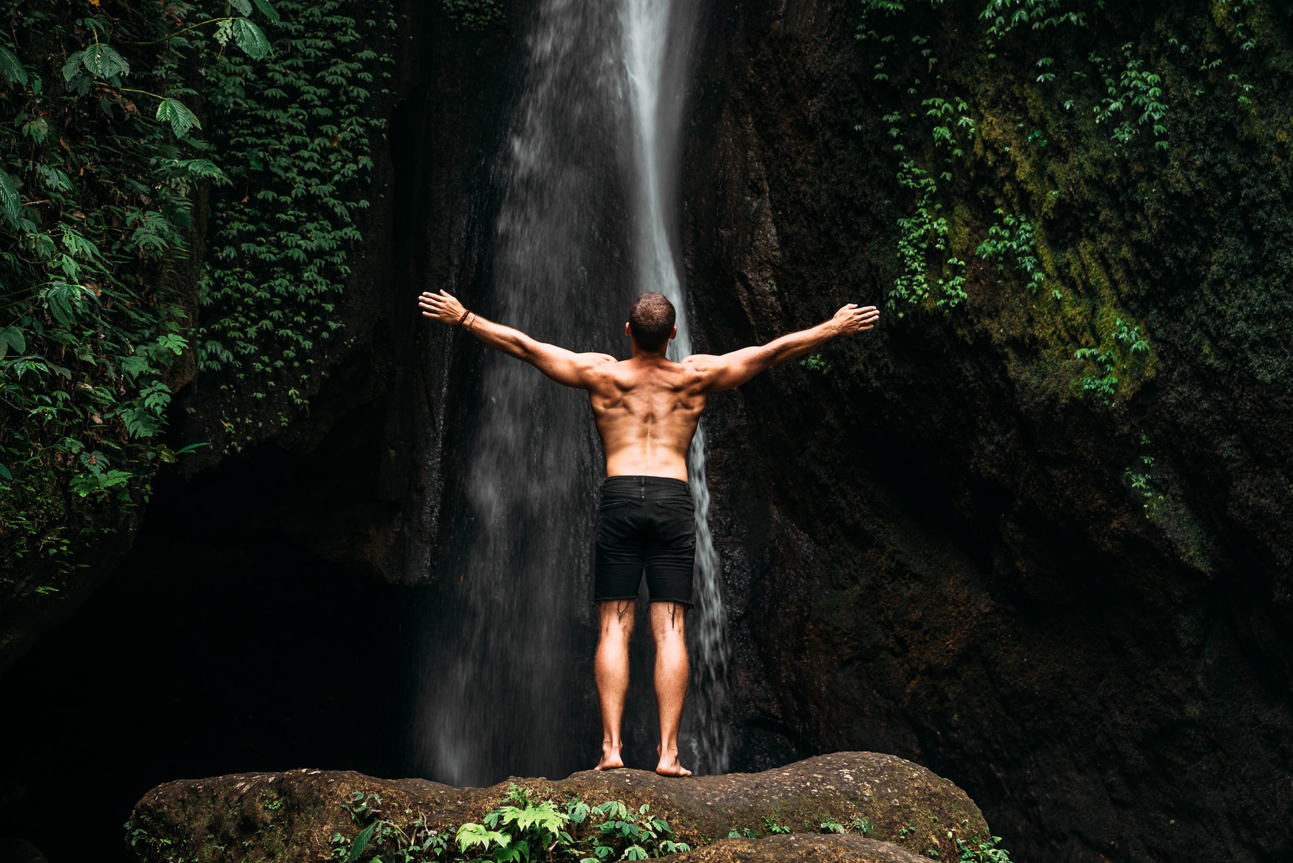 Back view of a man standing by waterfalls with arms outstretched. Man at waterfall raising his hands in feeling closer to nature. Man at the waterfall. Travel to Bali. Man on the background of nature